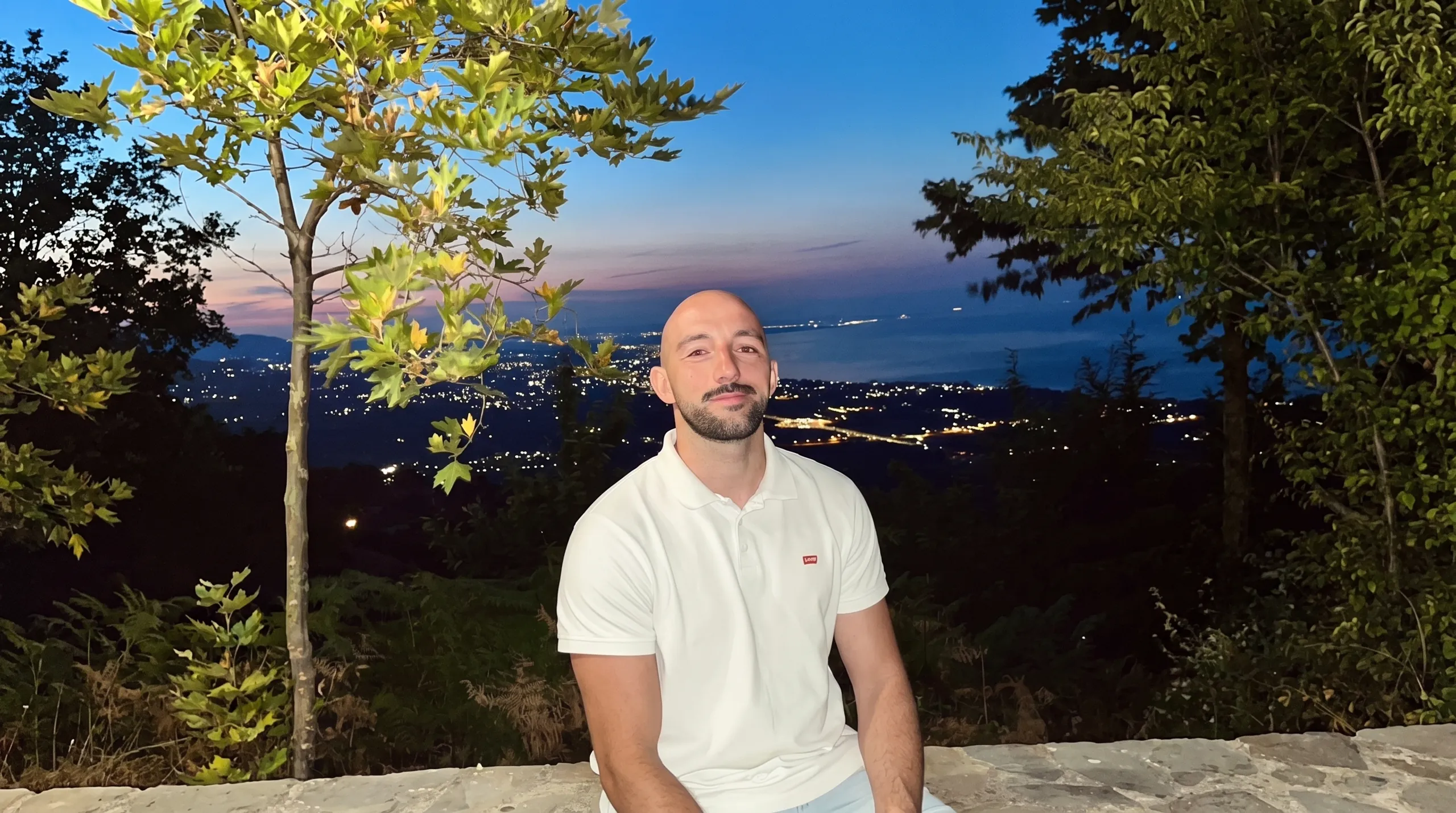 Sotiris at dusk above Palaios Panteleimonas, Pieria, with the lights of the plain and the sea behind him